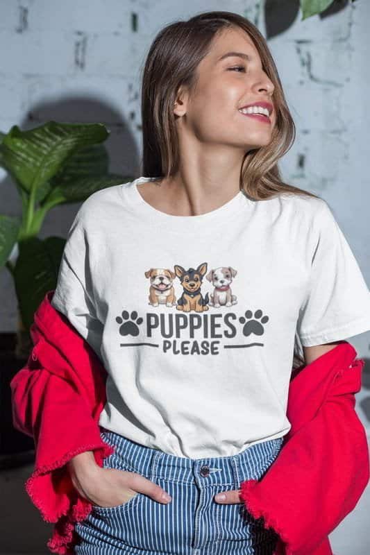 A cheerful young woman wearing a white t-shirt with a cute "Puppies Please" design featuring three illustrated puppies and paw prints. She pairs it with striped jeans and a red jacket draped over her shoulders.