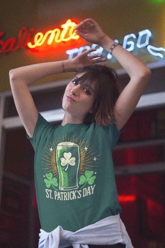 A young woman wearing a green St. Patrick’s Day t-shirt featuring a pint of green beer with a shamrock design and glowing rays. She poses playfully in front of neon lights, embracing a festive and fun vibe.