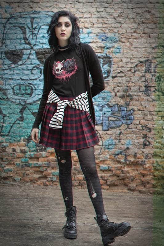A gothic-style woman wearing a black t-shirt with a pink artistic design of two gothic cats. She pairs it with a red plaid skirt, ripped tights, combat boots, and a black cardigan, posing in front of a graffiti-covered brick wall.