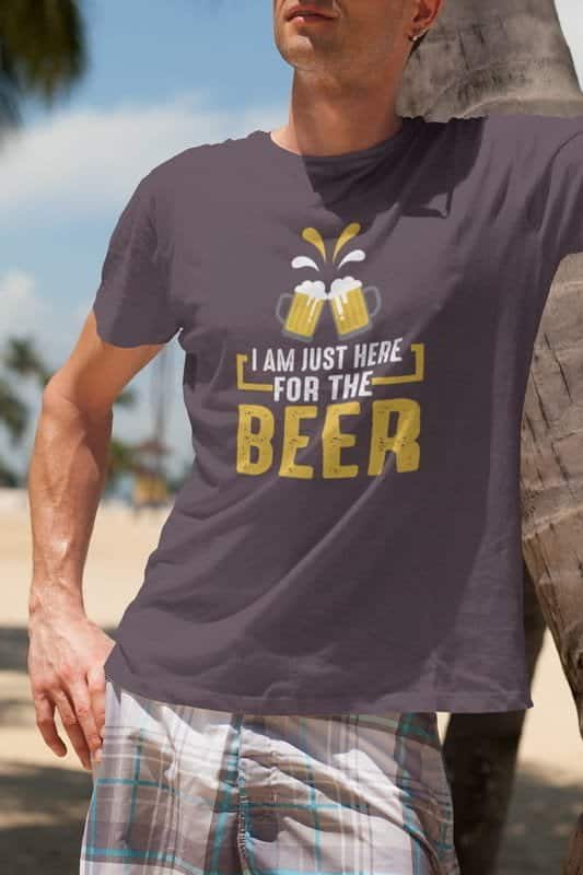 A relaxed man wearing a dark gray t-shirt with the phrase "I Am Just Here for the Beer" and an illustration of two clinking beer mugs. He is leaning against a palm tree on a sunny beach, wearing plaid shorts.